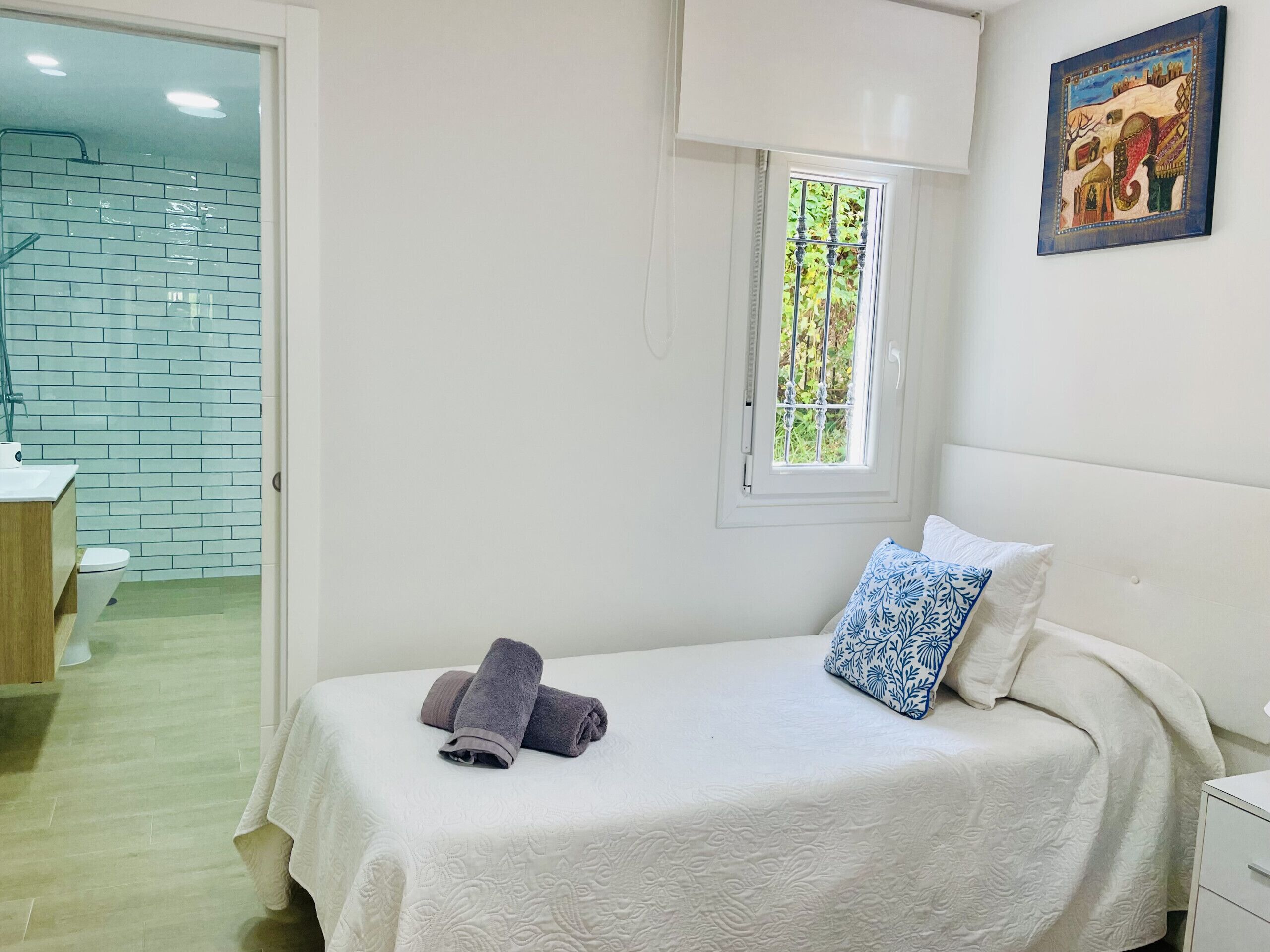 A bedroom with a bed, a window, a painting, and a bathroom visible in the background.