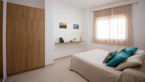 Un dormitorio con cama, armario, estante con libros y un florero, además de una ventana con cortinas.