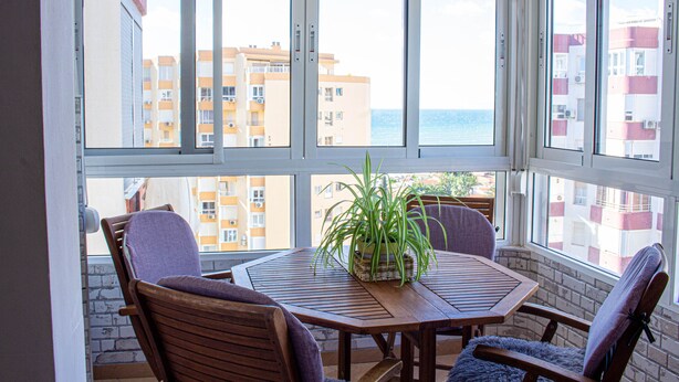 A room with a wooden table and chairs, a potted plant, and a view of buildings and the sea through large windows. - Apto. Pescamar by At Home Costa del Sol (Torrox)