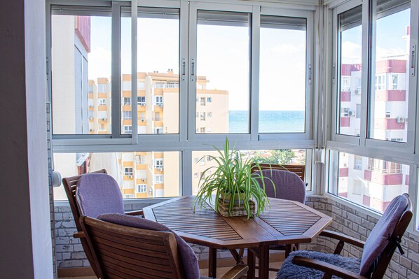 Ein Zimmer mit einem Holztisch und Stühlen, einer Topfpflanze und Blick auf Gebäude und das Meer durch große Fenster.