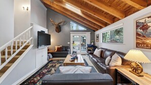 Un salon avec un plafond en bois, une tête de cerf montée au mur, un téléviseur, un canapé, une table basse et une lampe dont l’abat-jour est décoré d’un motif de roue de charrette.
