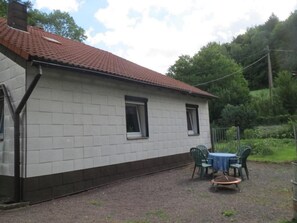 Ein eingeschossiges Haus mit einem Ziegeldach, einer kleinen Veranda mit Tisch und Stühlen sowie einer Auffahrt mit Kies.