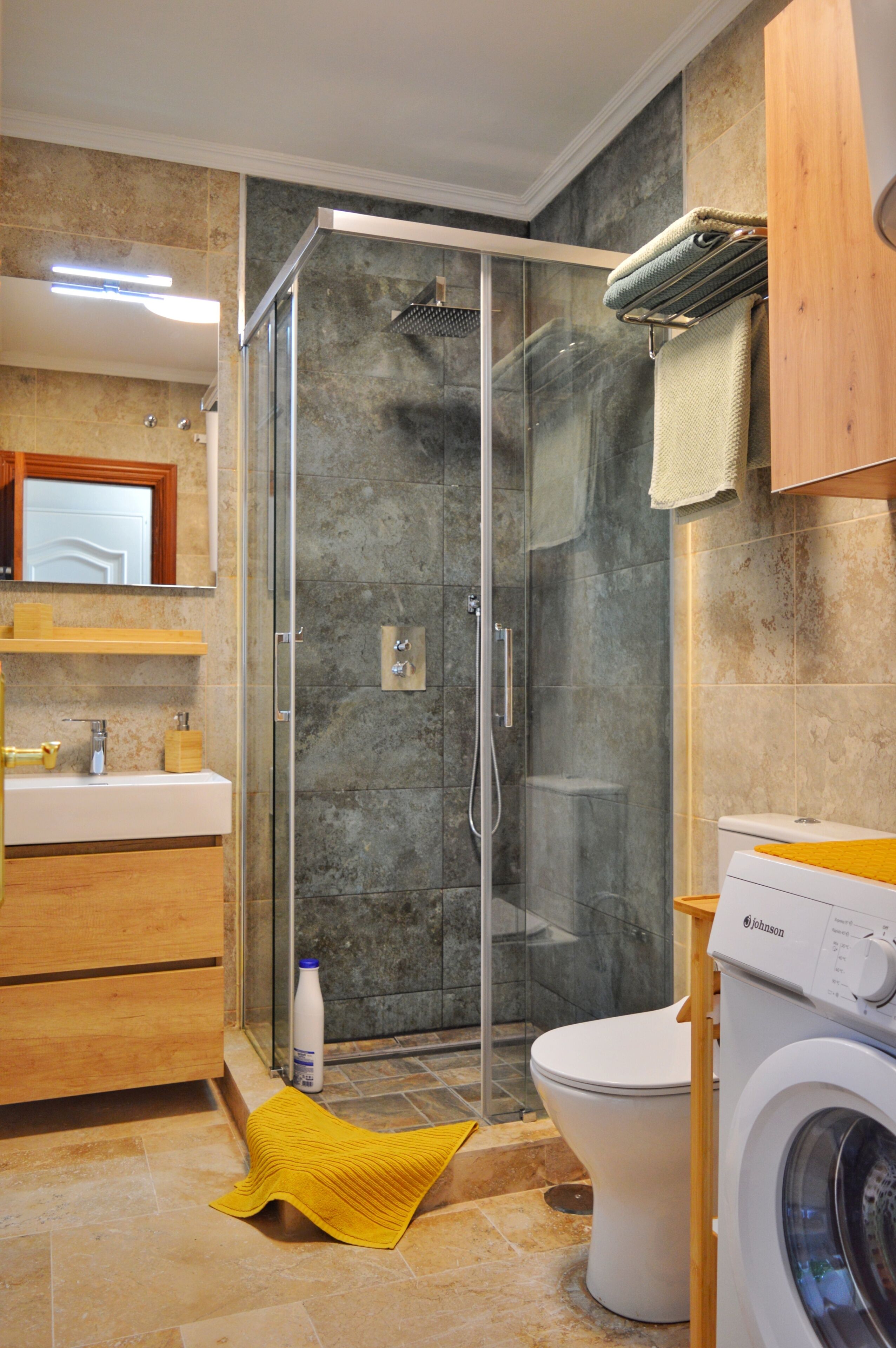 A modern bathroom with a glass-enclosed shower, a toilet, a washing machine, and a wooden vanity.