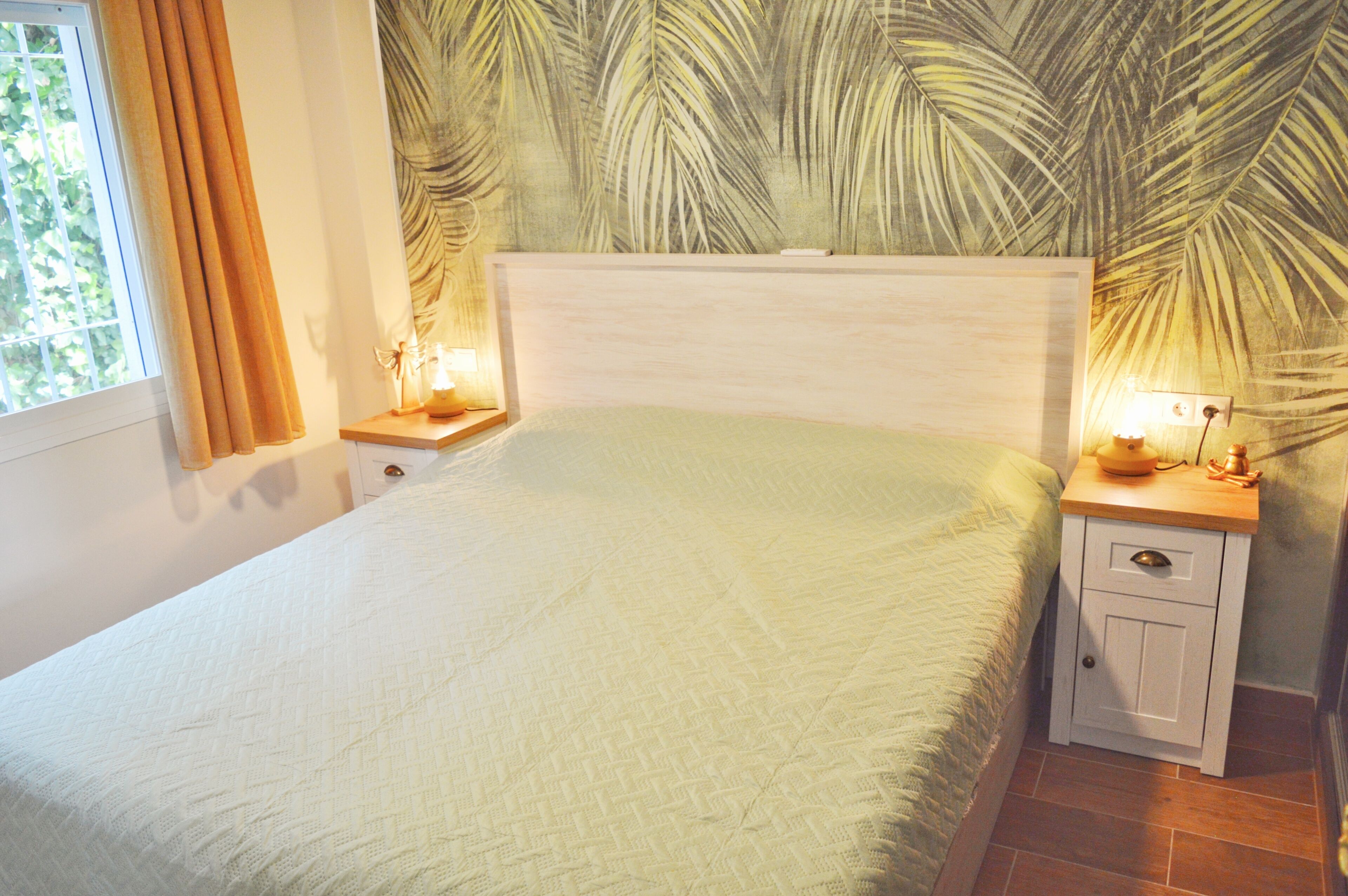 A bedroom with a bed, two bedside tables, a nightstand, a wall with a leaf pattern, and a window with curtains.