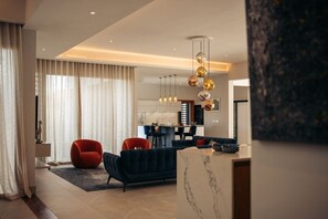 A modern living room with a dark sofa, red chair, and a marble coffee table. A kitchen with a bar and pendant lighting is visible in the background. - Designer Villa – Anahita Golf Resort Luxury Residence (Beau Champ)