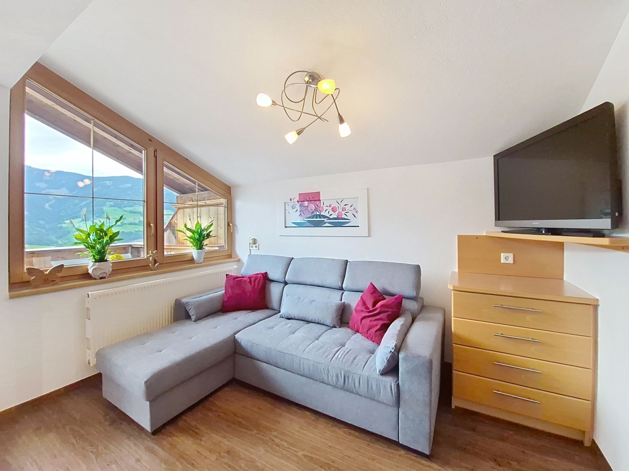 A modern living room with a grey sectional sofa, a wooden cabinet, a flat-screen TV, a ceiling light fixture, and a window with a view of the outdoors.