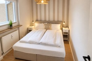 En pænt redt seng med hvide sengetøj, to sengeborde med lamper, en radiator, et vindue med udsigt til en murstensmur, og en stribet væg bag sengen.