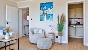 A living room with a sofa, a coffee table, a chair, a painting, a lamp, a vase with dried plants, and a kitchen area with cabinets and appliances. - Dünenglück No. 4 - The vacation apartment Dünenglück No. 4 on the beach for max. 4 (Sylt)