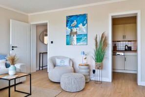 Ein Wohnzimmer mit einer Couch, einem Couchtisch, einem Sessel, einem Bild, einer Lampe, einer Vase mit Trockenpflanzen und einer Küchenzeile mit Schränken und Geräten.