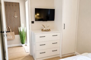 A bedroom with a bed, a white dresser with drawers, a television mounted above it, a wooden slat wall, and a plant. - Dünenglück No. 4 - The vacation apartment Dünenglück No. 4 on the beach for max. 4 (Sylt)