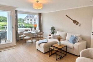En stue med sofa, sofabord, spisebord med stole og udsigt til en bygning gennem et vindue.