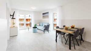A modern living room with a sofa, a dining table with chairs, a small round table with a plant, and a view of the outside. - MEERholung Vacation Rental 3.8:51 m² near the sea, sleeps up to 4, with a balcony - House (Cuxhaven)