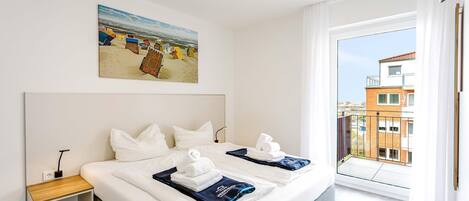 Habitación de hotel con una cama grande, una mesita de noche, un cuadro de cabañas en la playa y un balcón con vista a la ciudad.