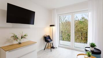 Ein modernes Wohnzimmer mit Flachbildfernseher, einem Holztisch, einem Sessel und einem Balkon mit Blick auf Bäume.