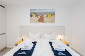Une chambre d’hôtel avec un lit, deux tables de chevet, un tableau représentant des chaises de plage accroché au mur, et une porte blanche.