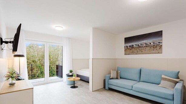A modern living room with a light blue sofa, a small round table, and a TV mounted on the wall. - MEERholung Vacation Rental 3.6 with Balcony for up to 5 Guests, 400 m from the Sea - House (Cuxhaven)