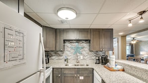 A kitchen with granite countertops, dark wood cabinets, a backsplash with a blue starfish design, and a map indicating room locations. - Walk to Ocean! Family PCB Condo w/ Balcony & Views (Panama City Beach)
