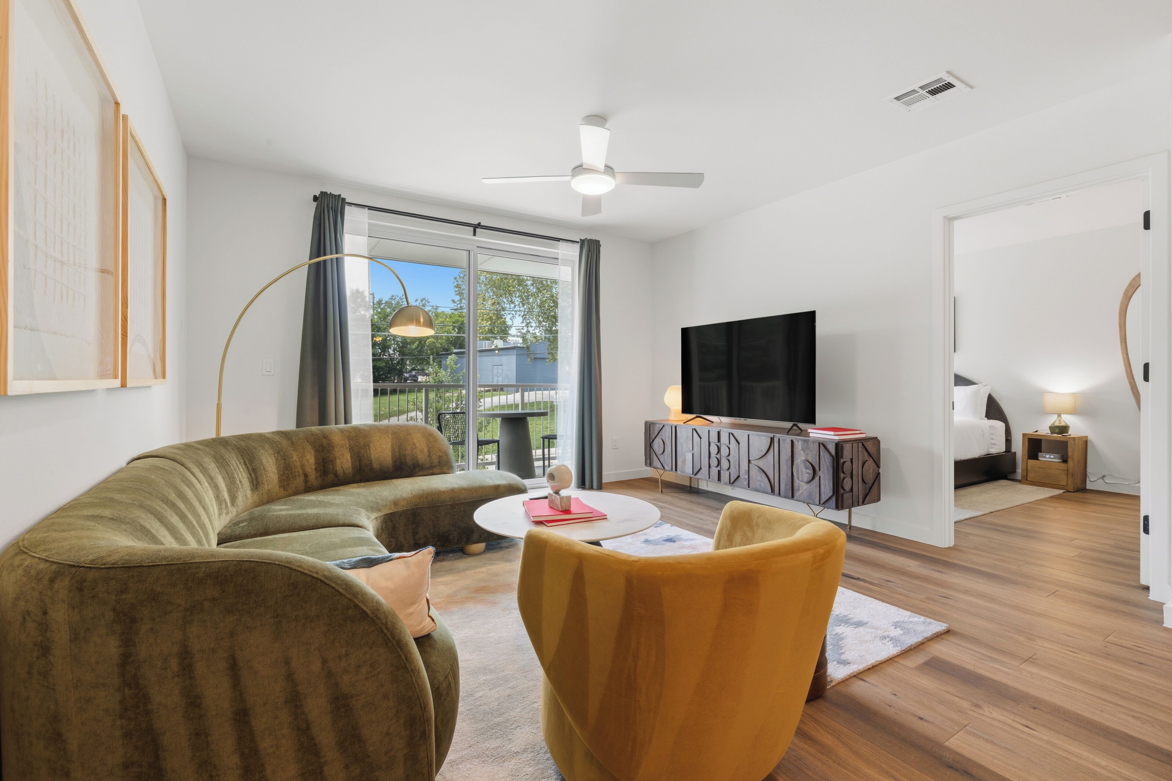 A modern living room with a sofa, armchair, and TV, a dining area with a table, and a balcony with a view.
