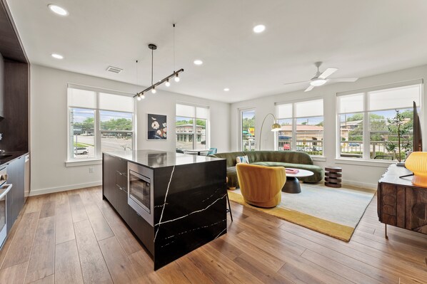 A modern kitchen with a black marble island, wooden flooring, and large windows offering city views.