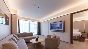Ein modernes Hotelzimmer mit einer großen Couch, einem runden Couchtisch, einem flachbildfernseher an der Wand und einem separaten Schlafbereich mit einem Bett.