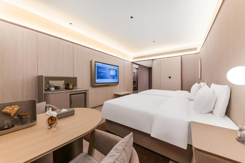 A modern hotel room with a large bed, a desk with a flat-screen TV, a small kitchenette, and a round dining table.