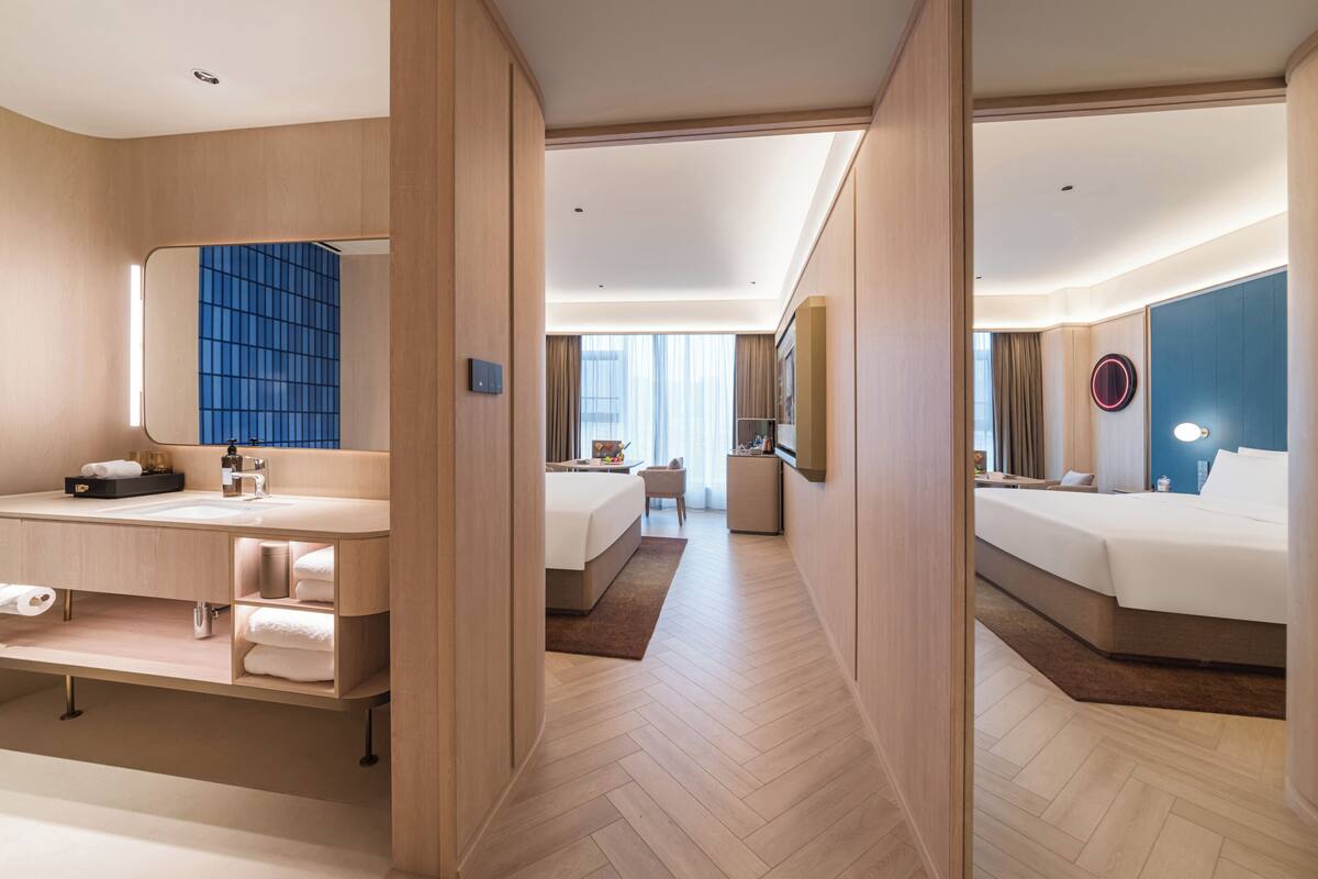 A hotel room with a large mirror, a vanity with a sink, a bed, a small desk, and a view of the city.