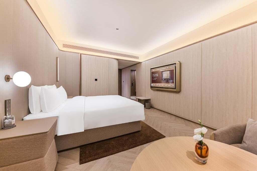 A modern hotel room with a bed, bedside table, TV, and a small table with a vase.