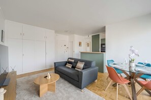 Ein modernes Wohnzimmer mit grauem Sofa, einem runden Couchtisch und einem Essbereich mit Glastisch und orangefarbenen Stühlen.