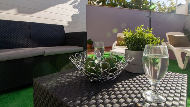 Eine Terrasse mit einem gewobenen Tisch, einer Metallschale, einem Glas Wasser, einer Couch und einem Blumentopf.