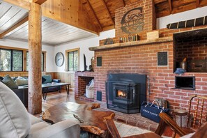A cozy living room with a fireplace, wooden furniture, and a large window. - Foosball + Firepit + BBQ | Near Lake + Slopes | Cherry Pine Chalet (Big Bear Lake)