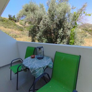 Uma varanda com duas cadeiras verdes, uma mesa pequena com toalha de mesa estampada, uma tigela vermelha e uma gaiola de pássaro preta.