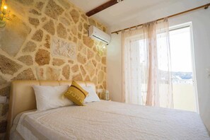 A bedroom with a stone wall, a bed with white and yellow pillows, and a view through a window with sheer curtains. - Farkia Exclusive Suites (Faliraki)