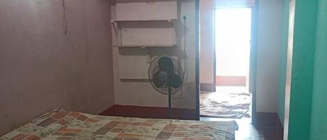 A bedroom with a bed, a ceiling fan, a standing fan, a window, and a door leading to another room.