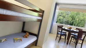 A bunk bed with a mattress, a teddy bear, and a children's book. There is a dining table with chairs and a view of trees through a window. - Nature Studio for 4 in Chames (Vallon-Pont-d’Arc)