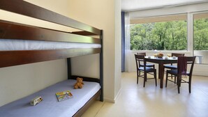 A bunk bed with a small toy car and a teddy bear on the lower bunk, a dining table with chairs, and a view of trees through large windows. - Nature Studio for 4 in Chames (Vallon-Pont-d’Arc)