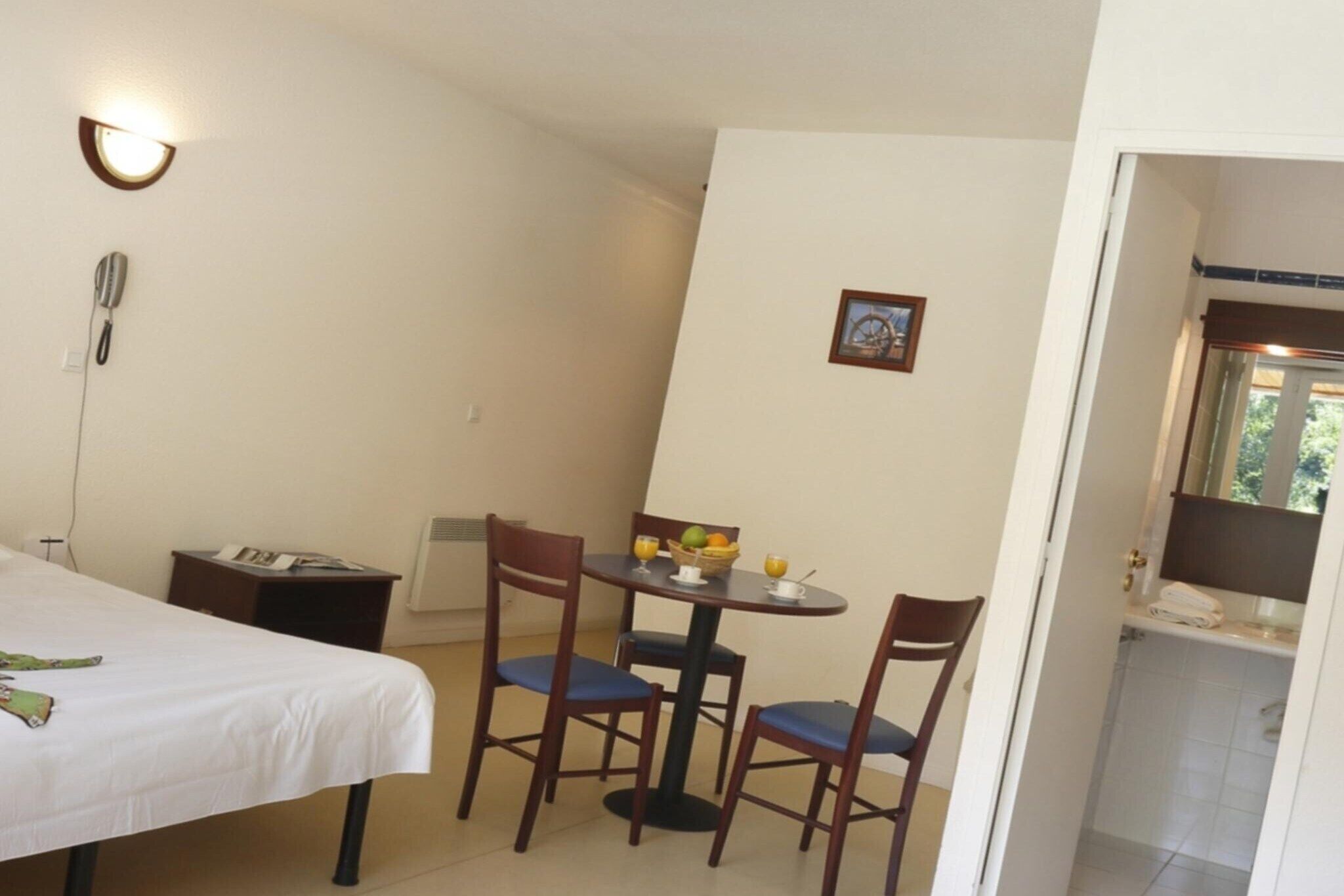 Une pièce comprenant un lit, un bureau avec un téléphone, une table avec des chaises, un panier de fruits et un tableau accroché au mur.