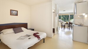A bedroom with a bed, a nightstand, a small table, a chair, a dining area with a table and chairs, and a kitchen area with a sink and cabinets. - Studio in the Heart of Nature (Vallon-Pont-d’Arc)