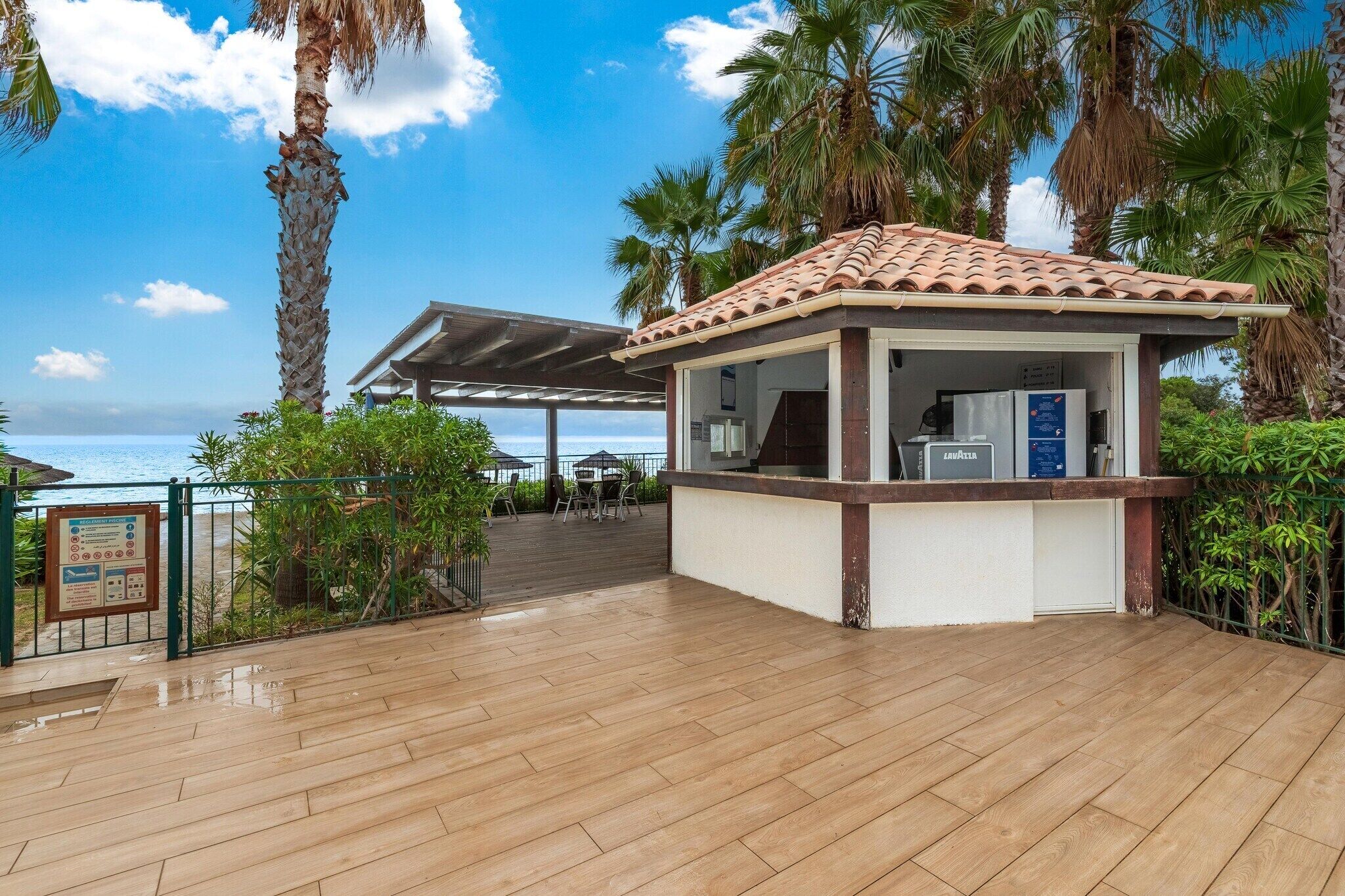 Een overdekt buitengebied met een houten terras, een klein kioskje met een pannendak en uitzicht op de oceaan.