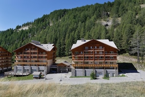 Un chalet composé de plusieurs bâtiments en bois, avec un télésiège visible au loin et un paysage montagneux et boisé en arrière-plan.