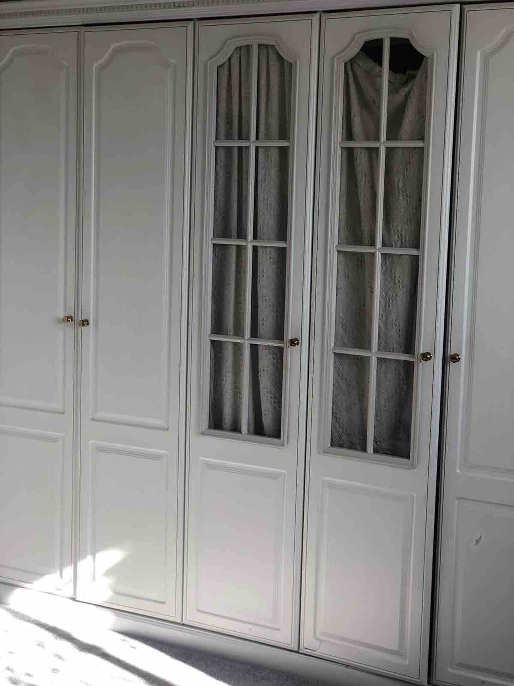 Ein weißer Kleiderschrank mit mehreren Türen und einem mittigen Fenster mit Vorhängen.
