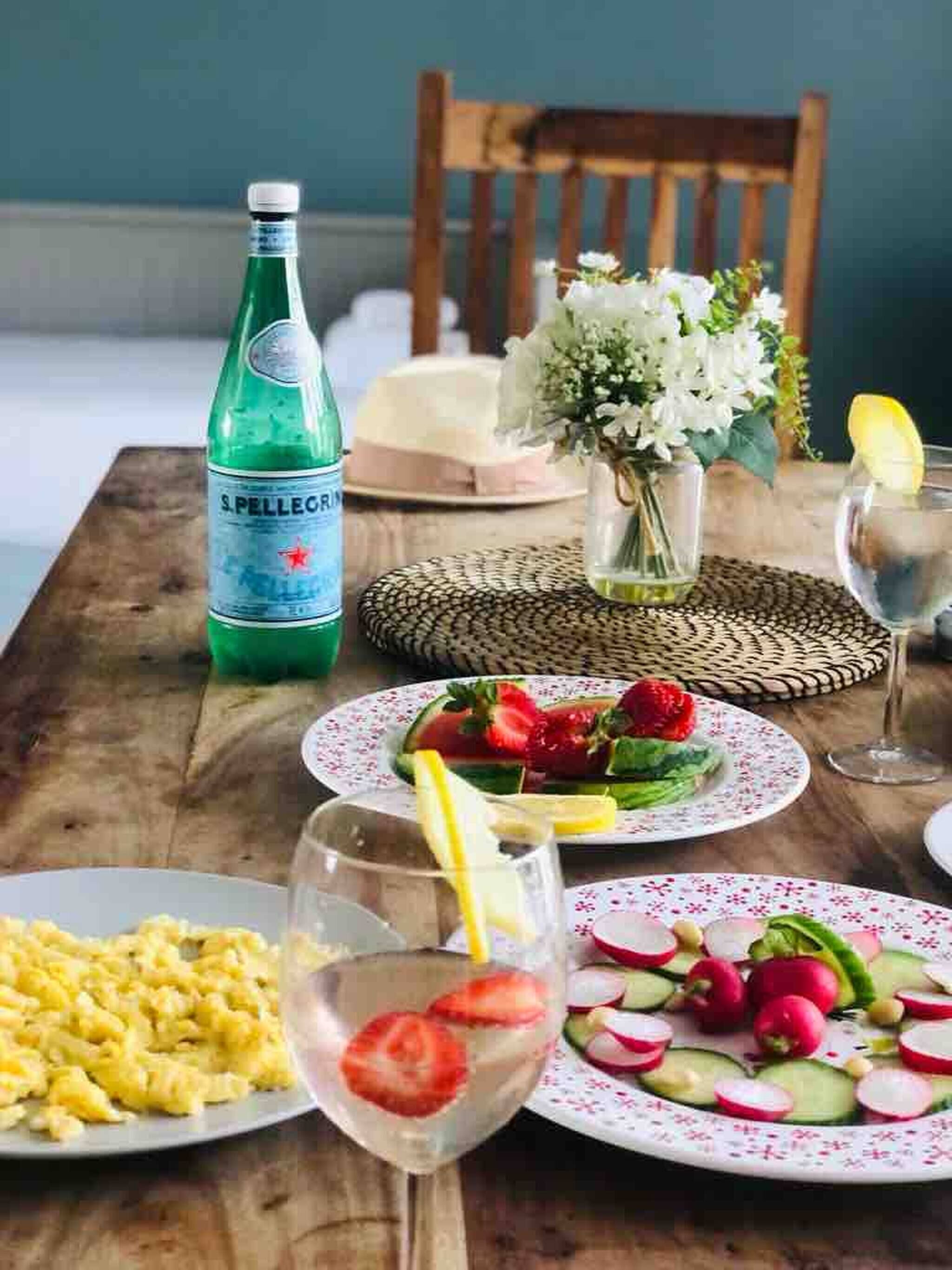 A wooden table with plates of food, a bottle of S. Pellegrino water, a vase with flowers, and a glass of water with a lemon slice.