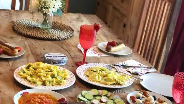 Een houten eettafel met borden eten, een bloemenvaas en een rood drankje.