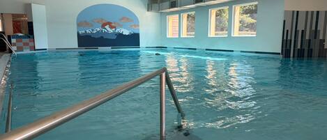 Piscina interior con vista a un mural de montaña, ventanas amplias y escalera metálica.