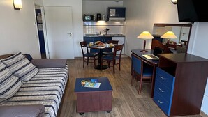 Un espacio habitable compacto con sofá, una pequeña mesa de comedor, kitchenette, escritorio con lámpara y televisor montado en la pared.