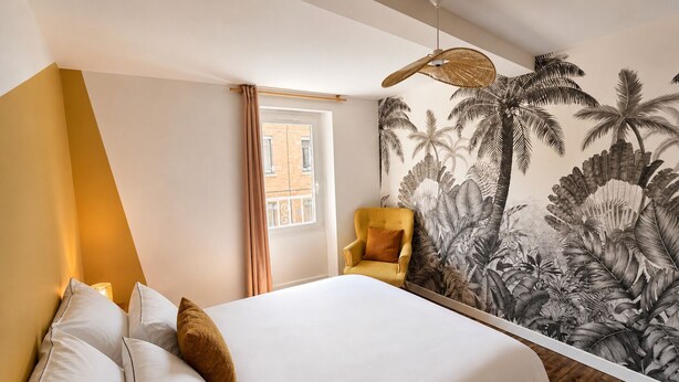 Un dormitorio con una cama, una silla, un ventanal y una pared con motivos de hojas tropicales.