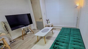 Ett vardagsrum med en platt-TV, en grön soffa, ett litet bord och ett fönster med persienner.