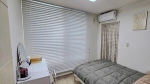 Ett sovrum med en säng, ett skrivbord med en stol, ett fönster med persienner, en luftkonditioneringsanläggning och en tavla som hänger på väggen.