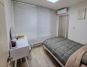 Ein Schlafzimmer mit Bett, Schreibtisch mit Stuhl, Fenster mit Jalousien, einer Klimaanlage und einem Wandbild.