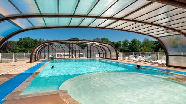 Una piscina techada con techo curvo, agua azul cristalina y sillones de descanso alrededor.