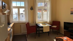 A compact living space with a kitchenette, two windows with curtains, a dining table, a red chair, a TV, and a wooden table. - Freundlich eingerichtete Wohnung mit Meerblick by Interhome (Dahme)